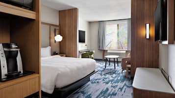 A hotel room with a bed, a desk, a chair, a coffee machine, a flat-screen TV, and a window with a view of trees.