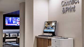 En receptionsdisk med en computerskærm, tastatur og printer. Et fjernsyn er synligt i baggrunden, der viser et nyhedsprogram.