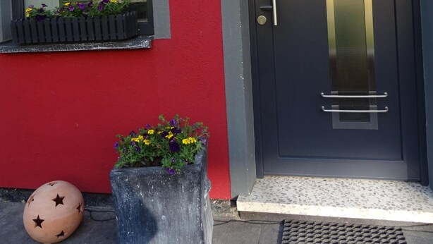 Una entrada de edificio con pared roja, puerta oscura con un letrero de bienvenida, ventana con vistas a una zona verde y maceta con flores.