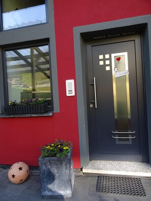 A building entrance with a red wall, a dark door with a welcome sign, a window with a view of a green area, and a planter with flowers. - Apartment newly renovated, brand new, very close to nature, fantastic location, (Ulmen -Meiserich)