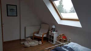 A room with a sloped ceiling, a radiator, a chair, a small table with a lamp, a toy house, and a large stuffed animal on the floor.