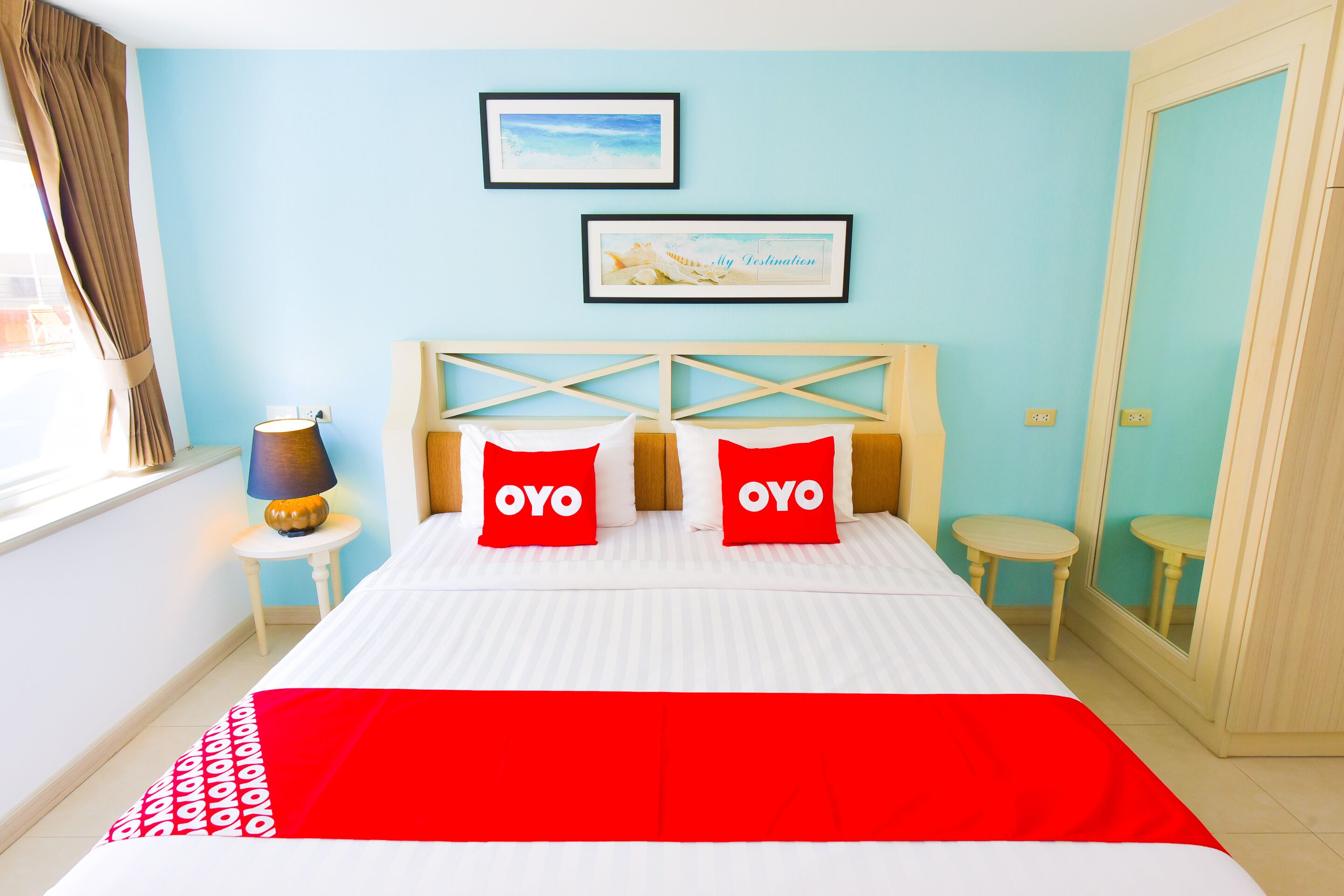 Ein Hotelzimmer mit einem Bett, zwei mit "OYO" markierten Kissen, einer Lampe und zwei gerahmten Bildern an der Wand.