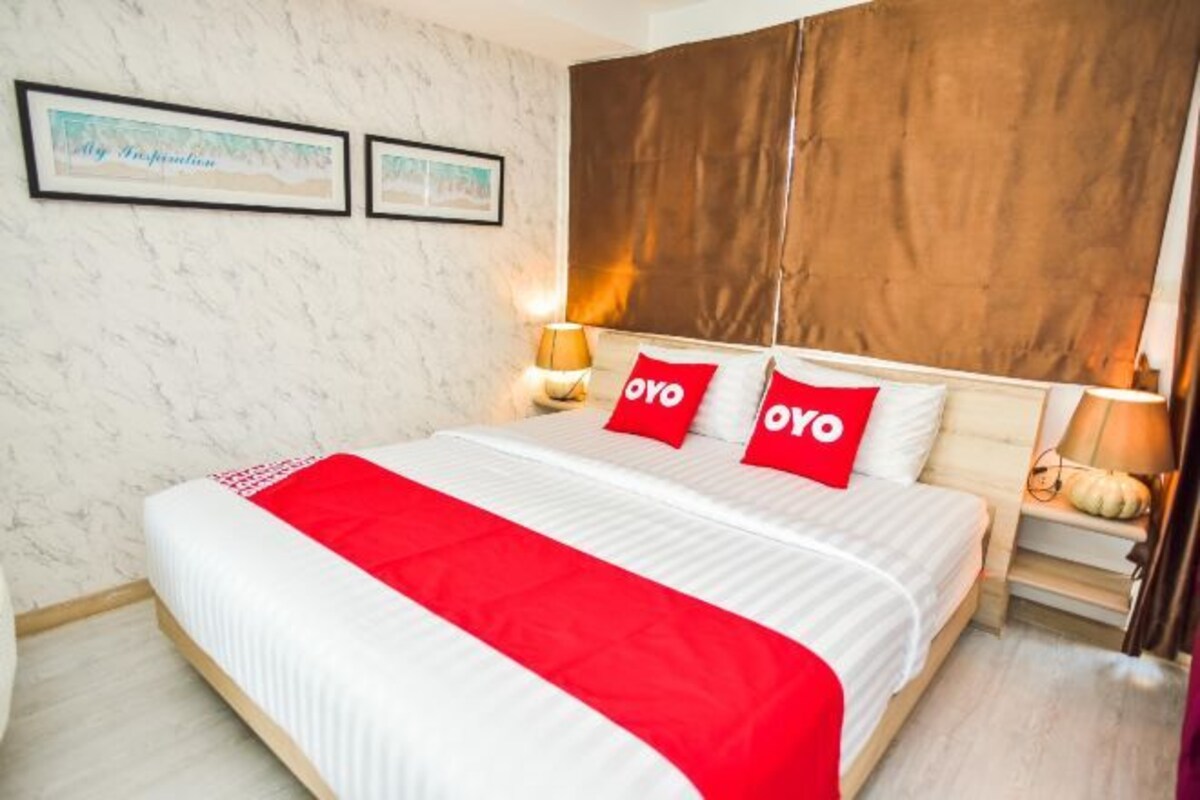 Hotellihuone, jossa on suuri sänky, kaksi «OYO»-logolla varustettua tyynyä, yöpöytä lampun kanssa ja seinällä olevia kehystettyjä kuvia.