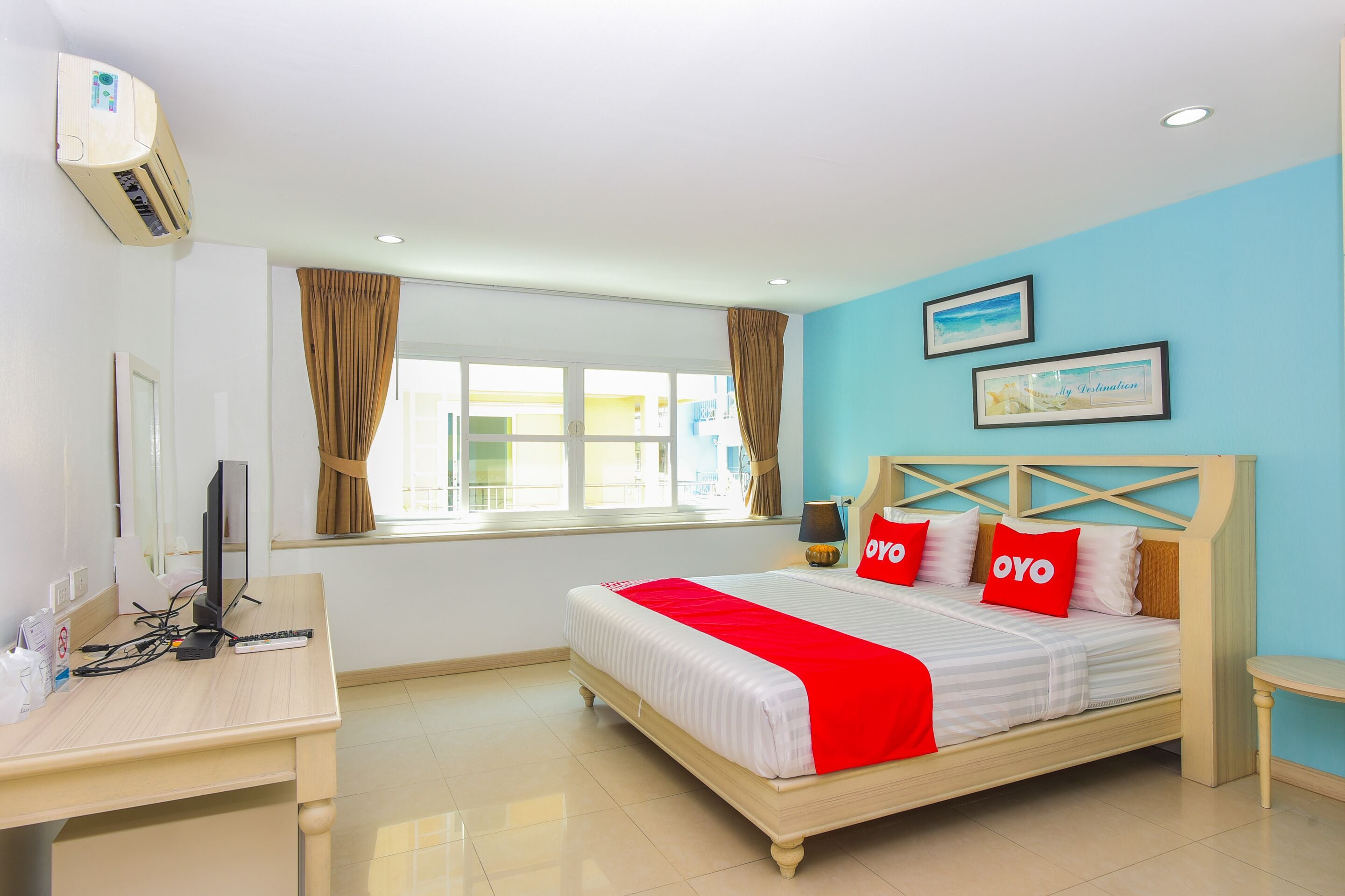 A hotel room with a bed, a desk, an air conditioner, a window with curtains, and a wall with framed pictures.