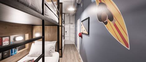 A bunk bed room with a wooden floor, a wall-mounted shelf with books, and a surfboard decoration.