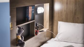 A neatly made bed with a white pillow and a wooden headboard. A small shelf holds a book titled "Australia" and a stuffed animal. A wall-mounted light illuminates the shelf area.