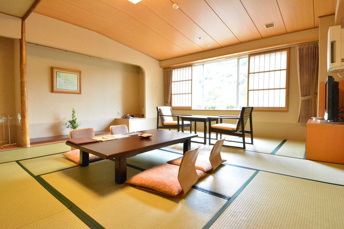 Ein traditioneller japanischer Raum mit Tatami-Böden, einem niedrigen Tisch, Stühlen und Blick ins Freie durch große Fenster.