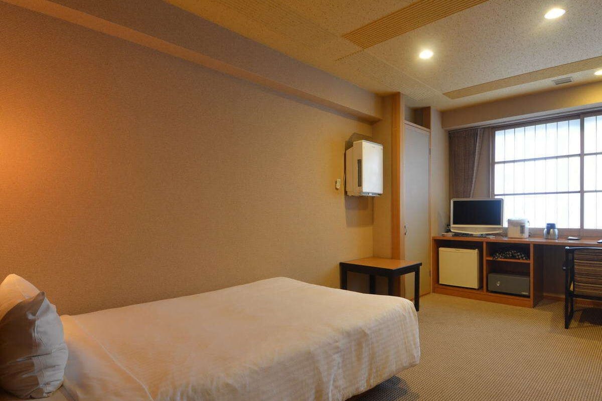 Habitació d'hotel amb un llit individual, una taula de nit amb televisió, un petit armari i una finestra amb cortines.