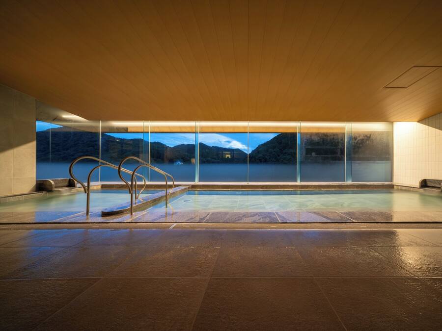 A large indoor swimming pool with a view of a lake and mountains, surrounded by tiled walls and a wooden ceiling.