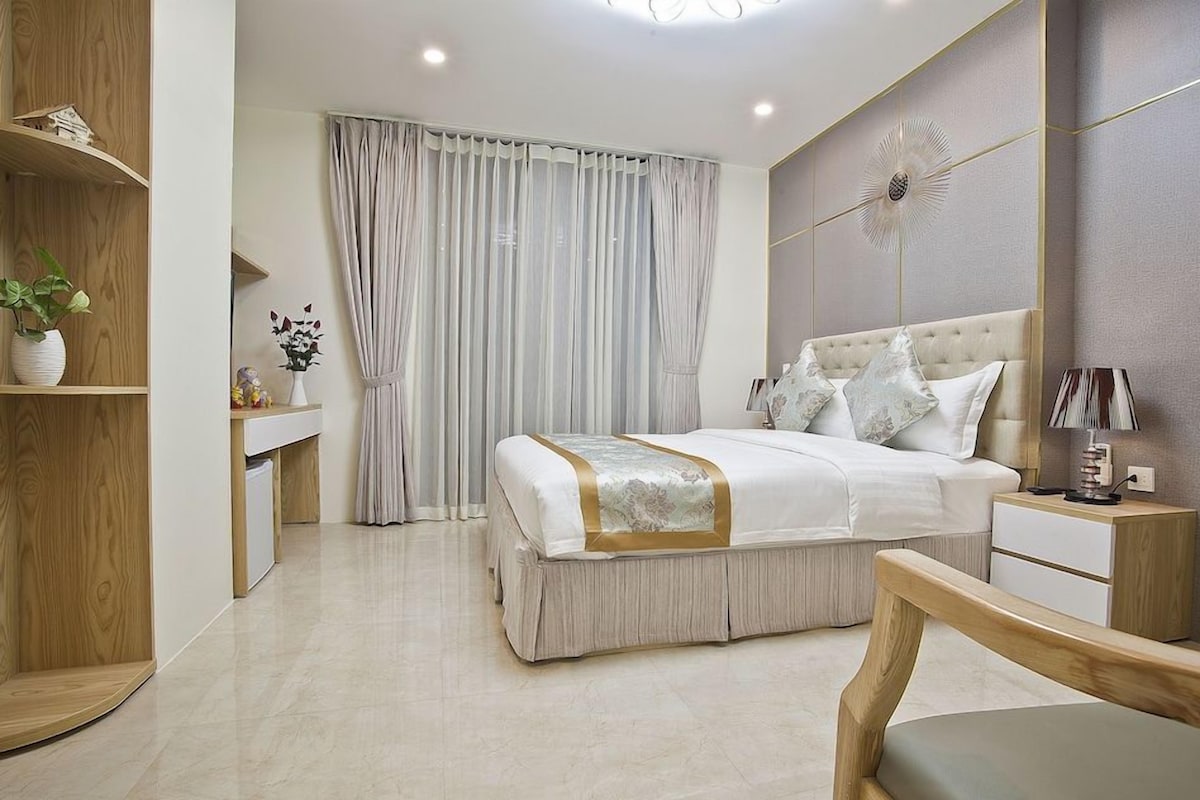 A modern bedroom with a large bed, bedside tables, a bedside lamp, a wooden chair, and a small desk with a plant.