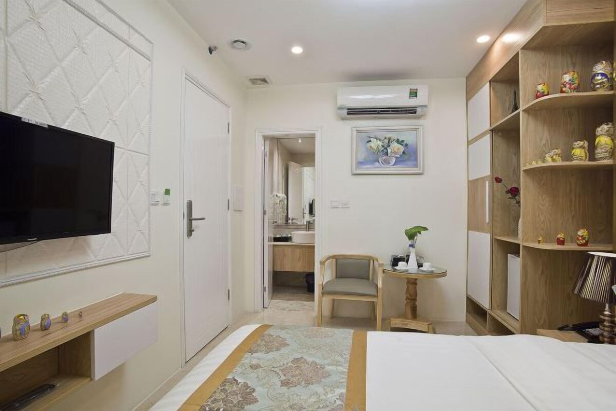 Une chambre d’hôtel comprenant un lit, une télévision, un bureau avec une chaise, une petite table avec une plante et une bibliothèque.
