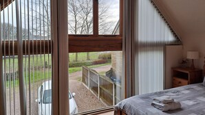 A bedroom with a large window, a bed with a blanket, a bedside table with a lamp, and a view of a car and garden outside. - Orchard Cottage (Stoke-on-Trent)