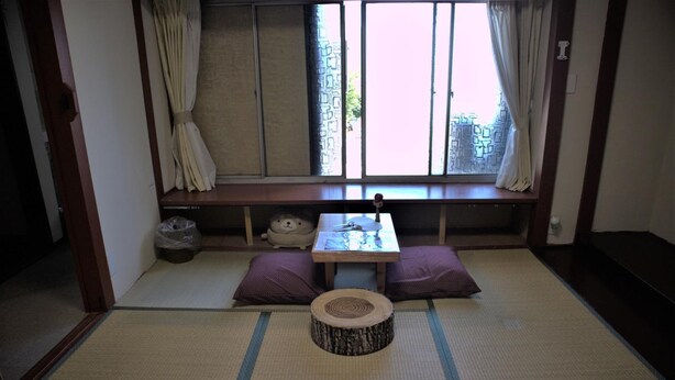 A traditional Japanese room with tatami flooring, a low table, cushions, and a window with curtains. - Hana Hostel Fujisan (Fujiyoshida)