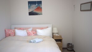 A neatly made bed with pink and white pillows, a wooden nightstand with a lamp, and a small trash can. - Hana Hostel Fujisan (Fujiyoshida)