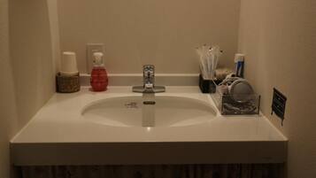 A bathroom sink with a white countertop, a red bottle, a white cup, and a small black cabinet.