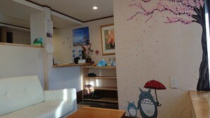A cozy room with a white sofa, a wooden table, and a wall decorated with a cherry blossom tree mural and a cat holding an umbrella. - Hana Hostel Fujisan (Fujiyoshida)