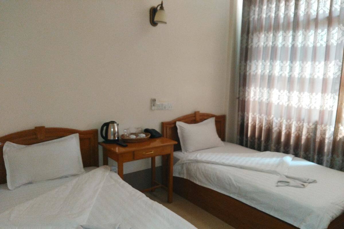 A hotel room with two single beds, a wooden nightstand with a kettle, a small table, and a wall-mounted lamp.