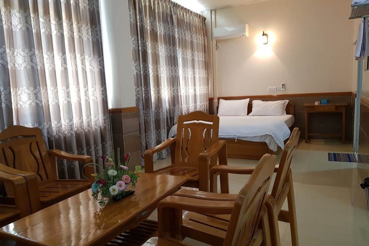 A hotel room with a wooden dining table, chairs, a bed with white bedding, a nightstand, and a wall-mounted air conditioning unit.