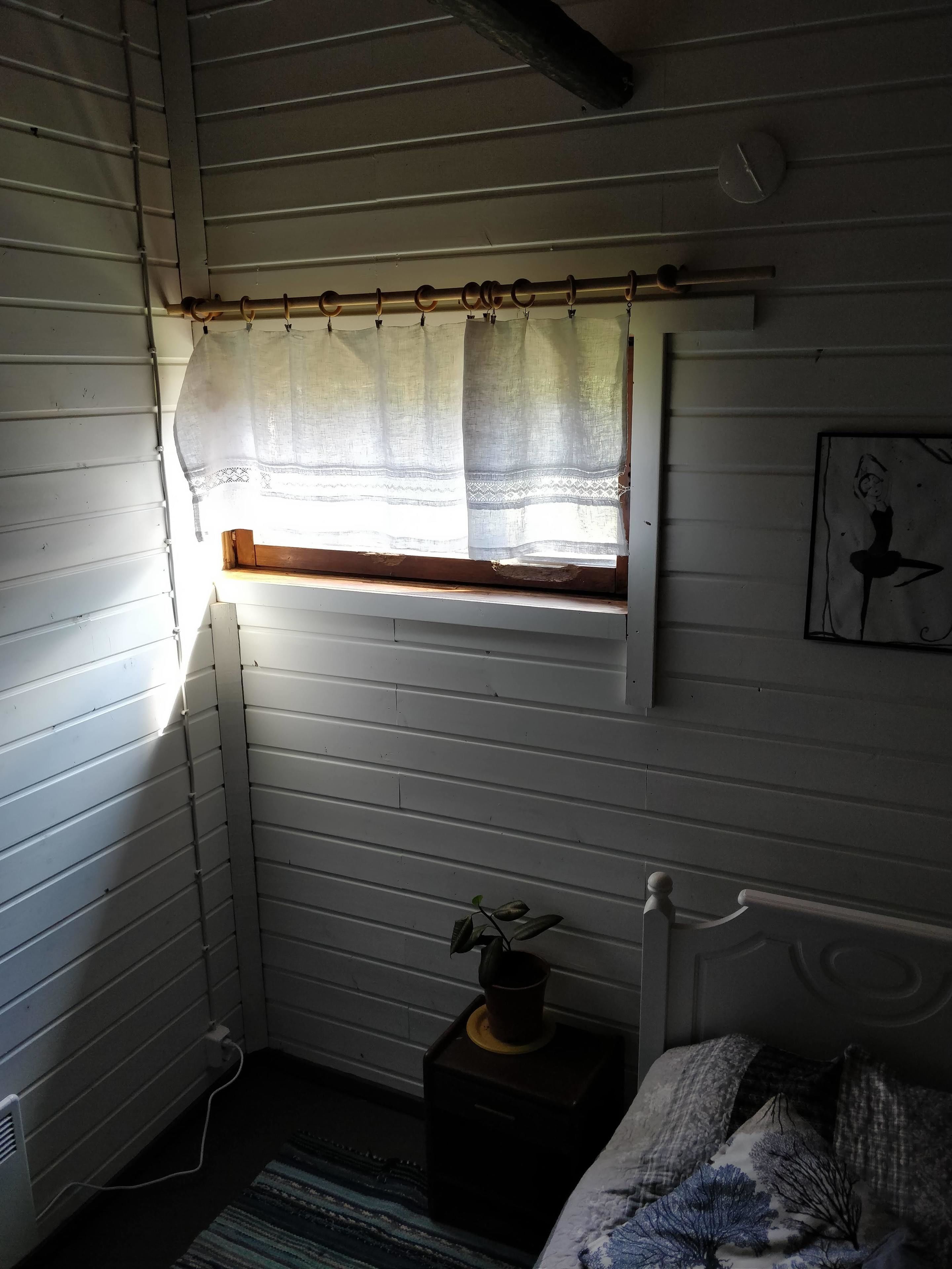 A room with a window covered by a sheer curtain, a radiator, a small plant on a wooden stand, and a bed with a patterned blanket.