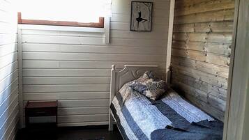 Una habitación pequeña con cama individual, techo y paredes de madera, una ventana con cortina, una mesita y un tapete azul a rayas.
