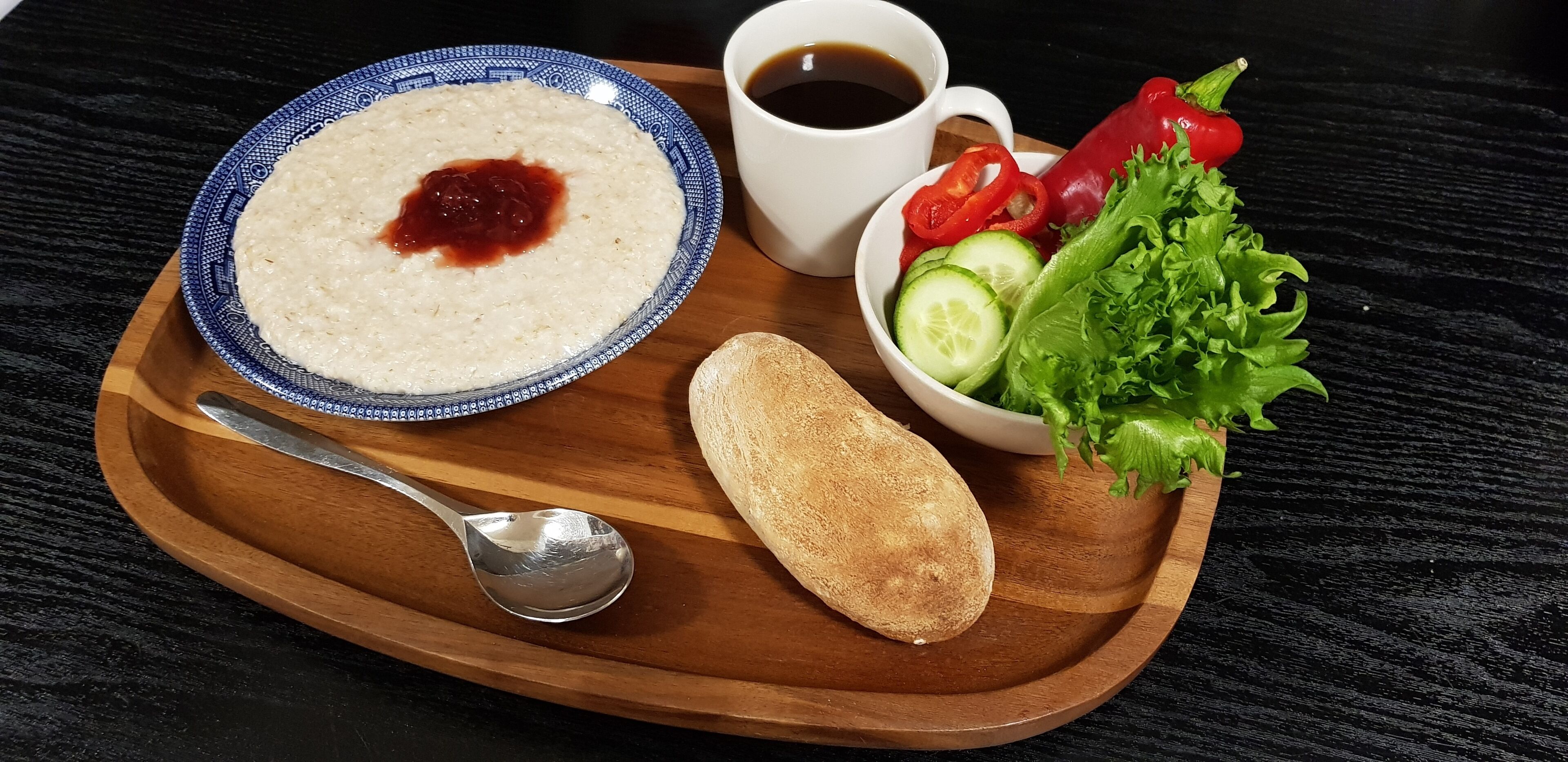 A wooden tray with a bowl of oatmeal, a cup of coffee, a loaf of bread, and a bowl of fresh vegetables.