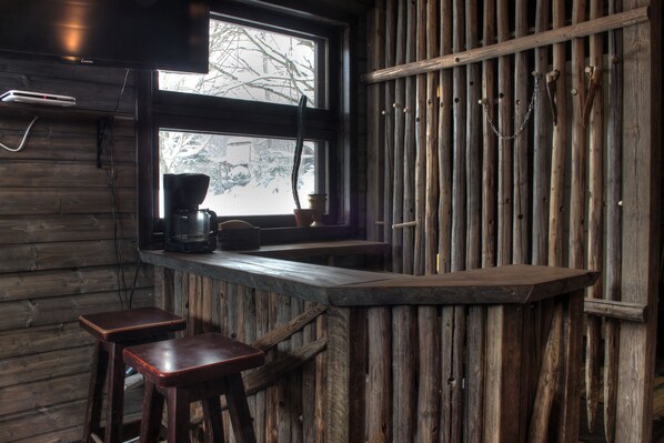 Un área de bar rústica de madera con una máquina de café, dos taburetes y un ventanal con vistas a árboles.