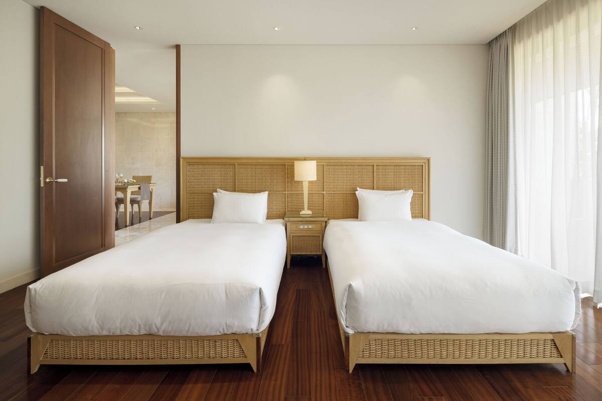 A hotel room with two single beds, a wooden headboard, a bedside table with a lamp, and a large window with curtains.