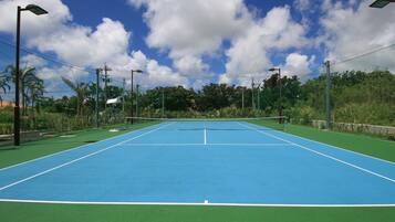 Una cancha de tenis al aire libre con superficies verdes y azules, rodeada de árboles y líneas eléctricas.