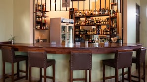 Zona de bar con un mostrador curvo, varias estanterías llenas de botellas, una vitrina refrigerada y tres taburetes de bar.