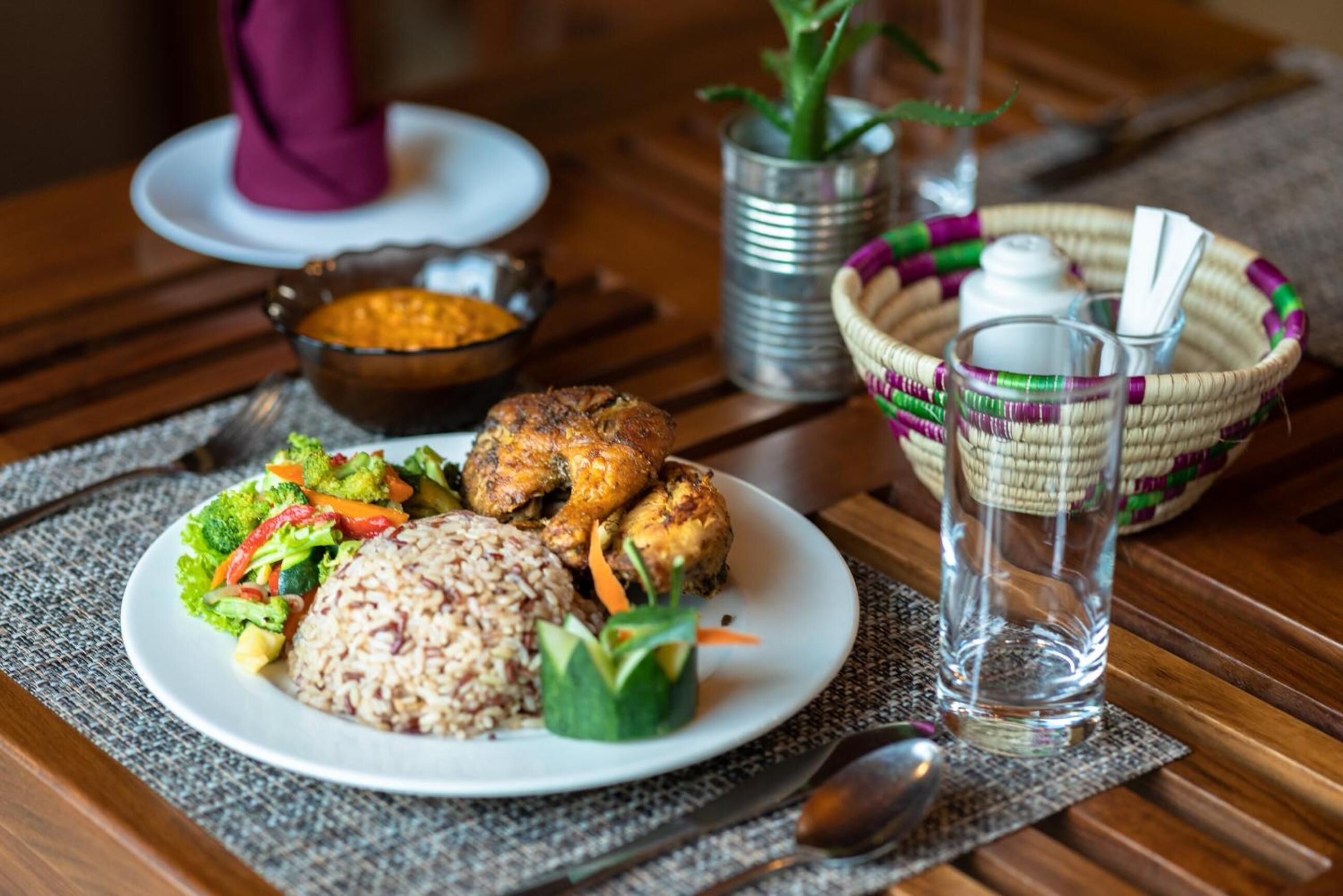 A plate of food with rice, vegetables, and chicken, a glass of water, and a woven basket with utensils on a wooden table.