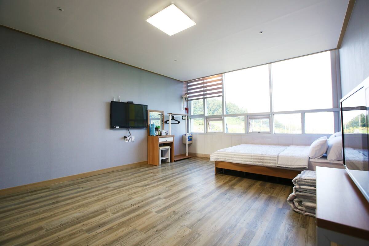 A modern bedroom with a wooden floor, a bed with white bedding, a desk with a computer, a TV mounted on the wall, and a window with blinds.