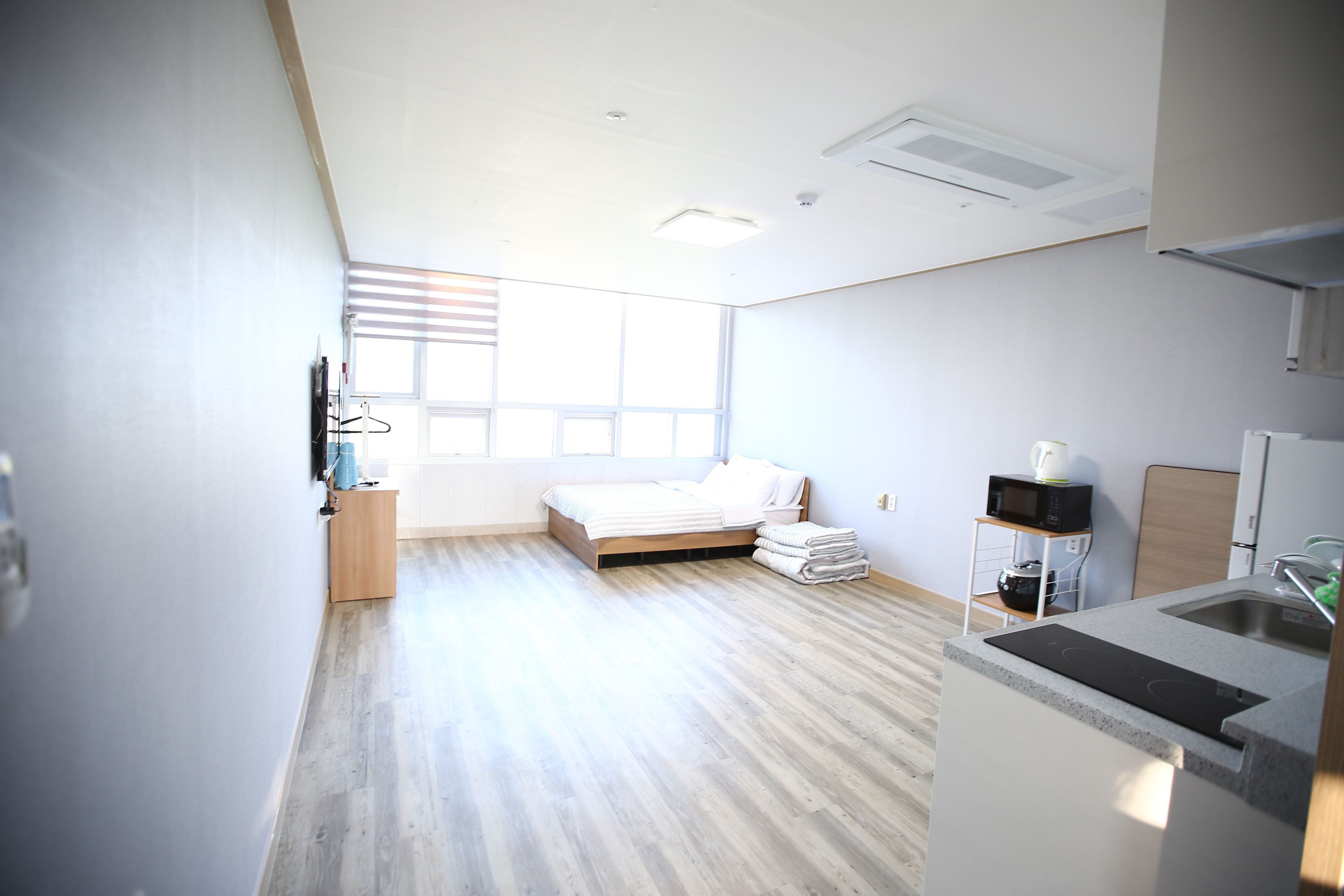A modern, well-lit bedroom with a wooden floor, a bed with white linens, a bedside table, and a window with blinds.