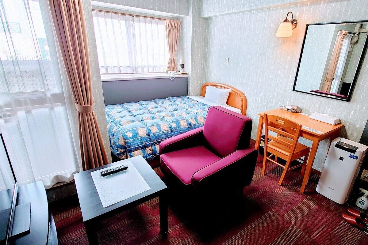 A hotel room with a bed, a desk, a chair, a small table, a mirror, a heater, and a window with curtains.