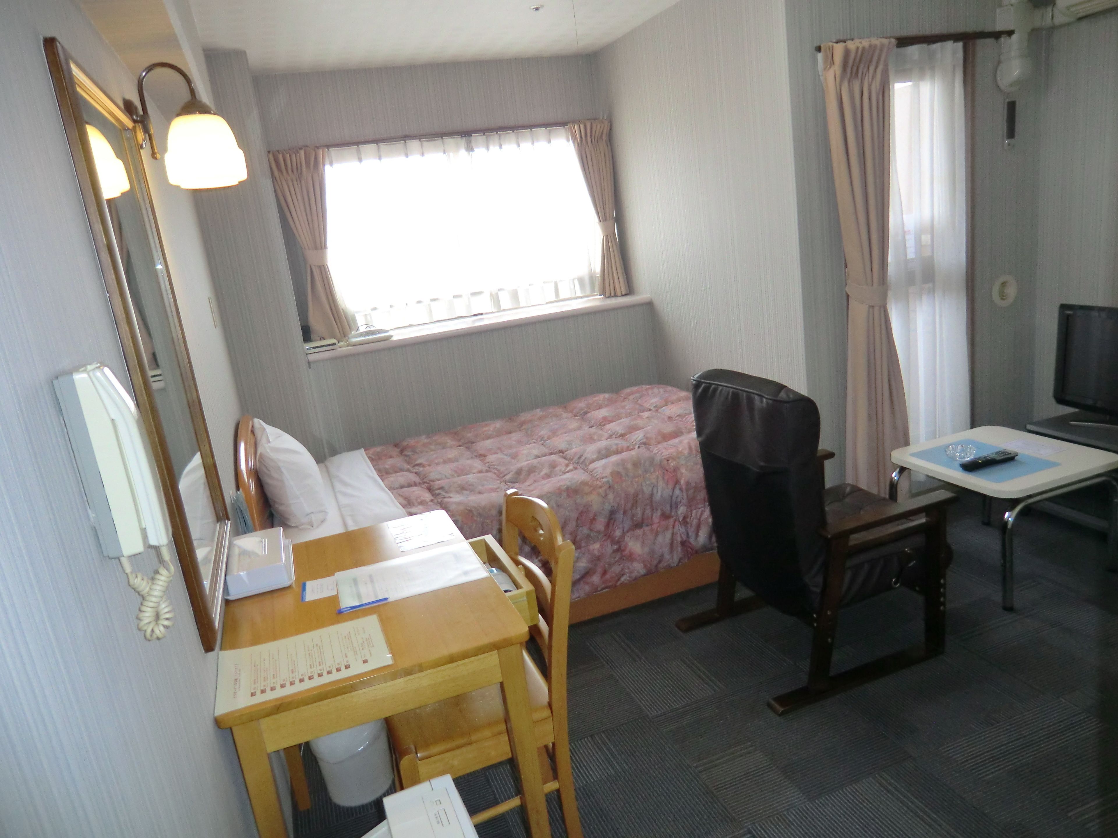 Una habitación de hotel con cama, escritorio, silla, televisor y ventana con cortinas.