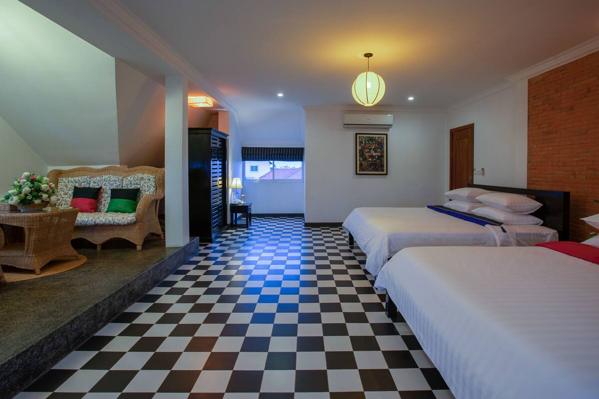 Habitación de hotel con piso a cuadros, dos camas, un sofá, una mesita y una puerta que da a otra habitación.
