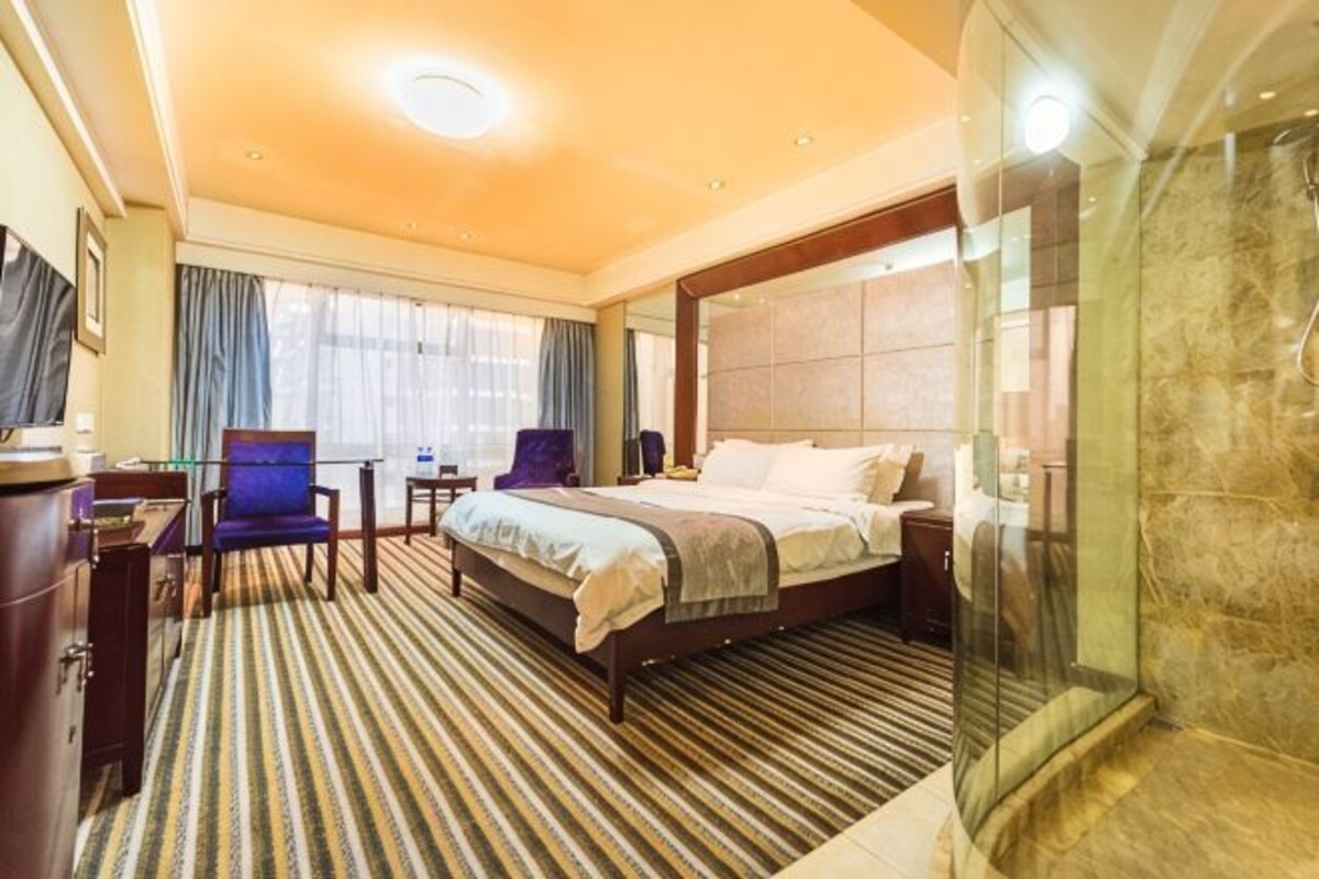 A hotel room with a large bed, a desk, a chair, a TV, and a shower area.