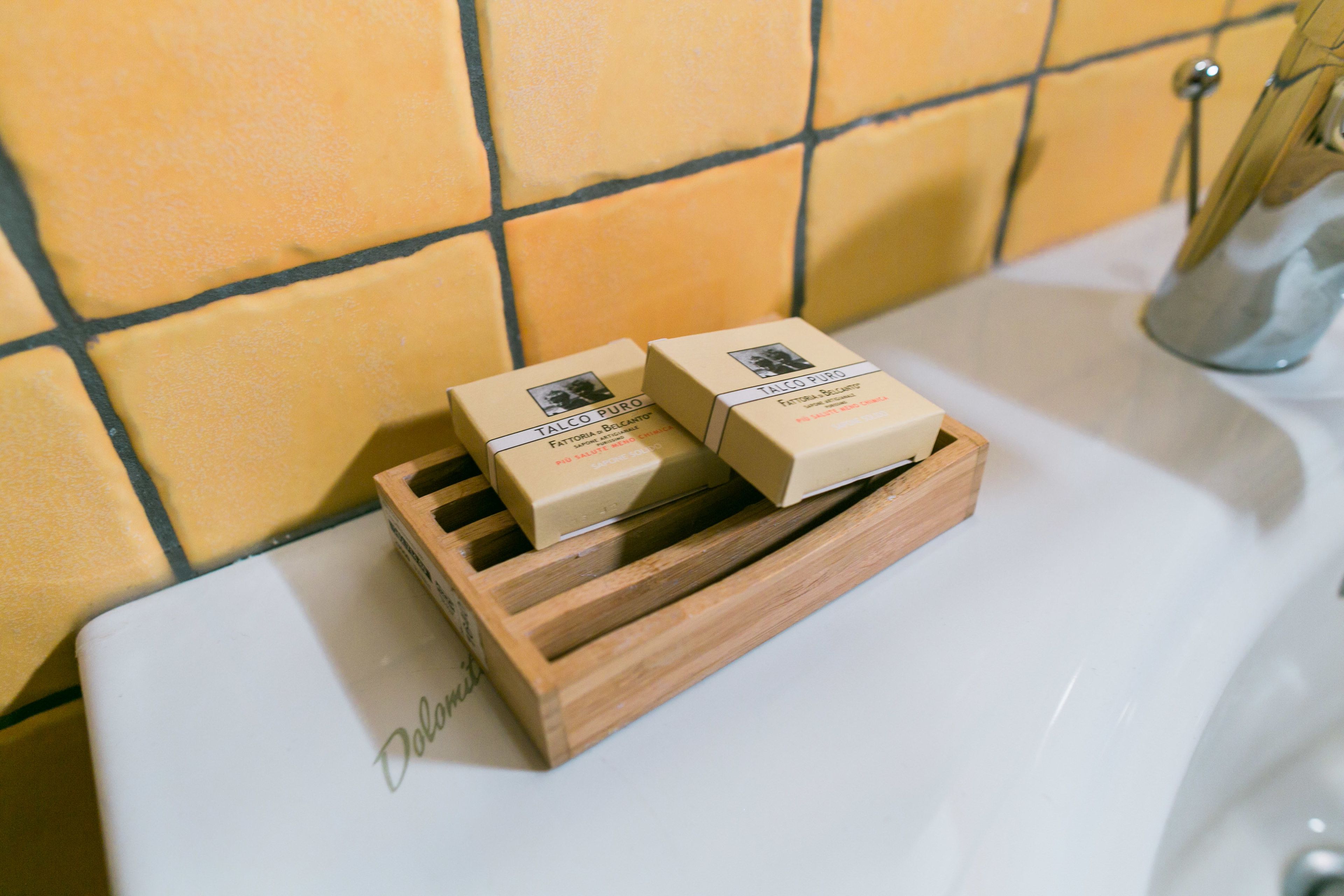 Dos pastillas de jabón en una bandeja de madera sobre un lavabo blanco con paredes de azulejos amarillos.