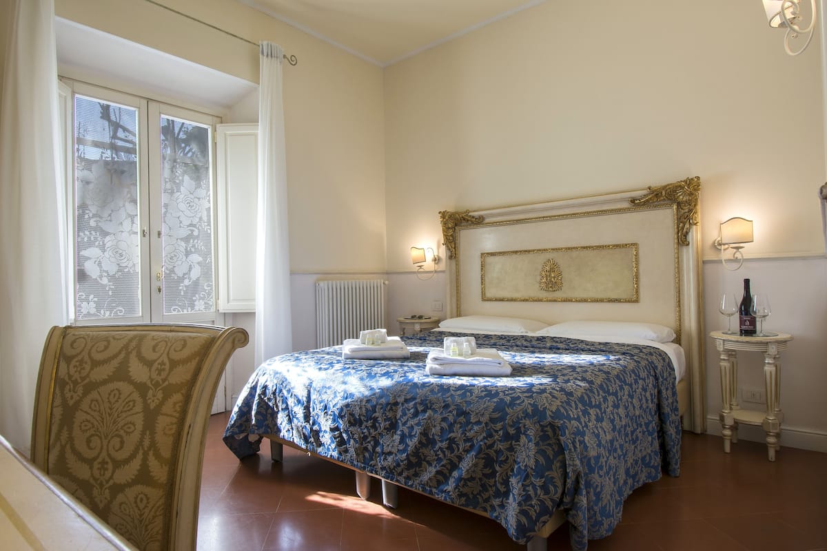 Habitación de hotel con una cama grande, una silla, una mesita de noche con lámpara y vistas al exterior a través de una ventana.