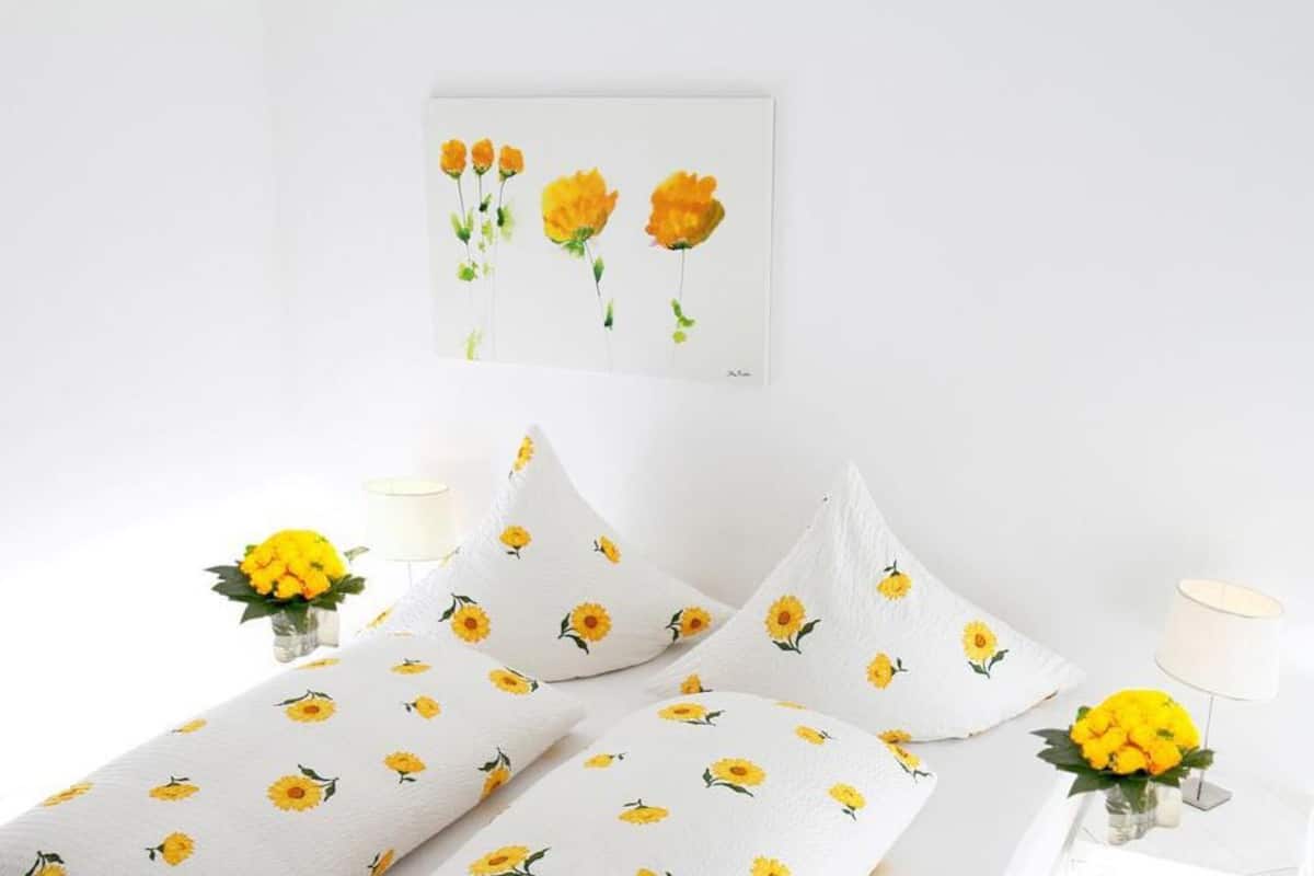 Ein Bett mit weißen Bettwäsche und Kissen, welche ein Sonnenblumenmuster zeigen, ein gerahmtes Sonnenblumenbild an der Wand und zwei Lampen auf beiden Seiten des Bettes.