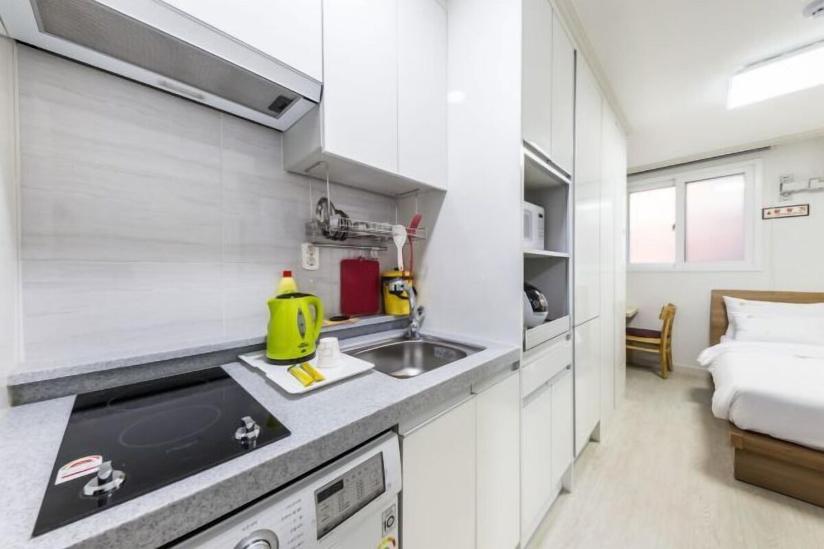 Una kitchenette compacta con estufa, microondas y lavamanos. En la habitación contigua hay una cama con ropa blanca y un cabecero de madera.