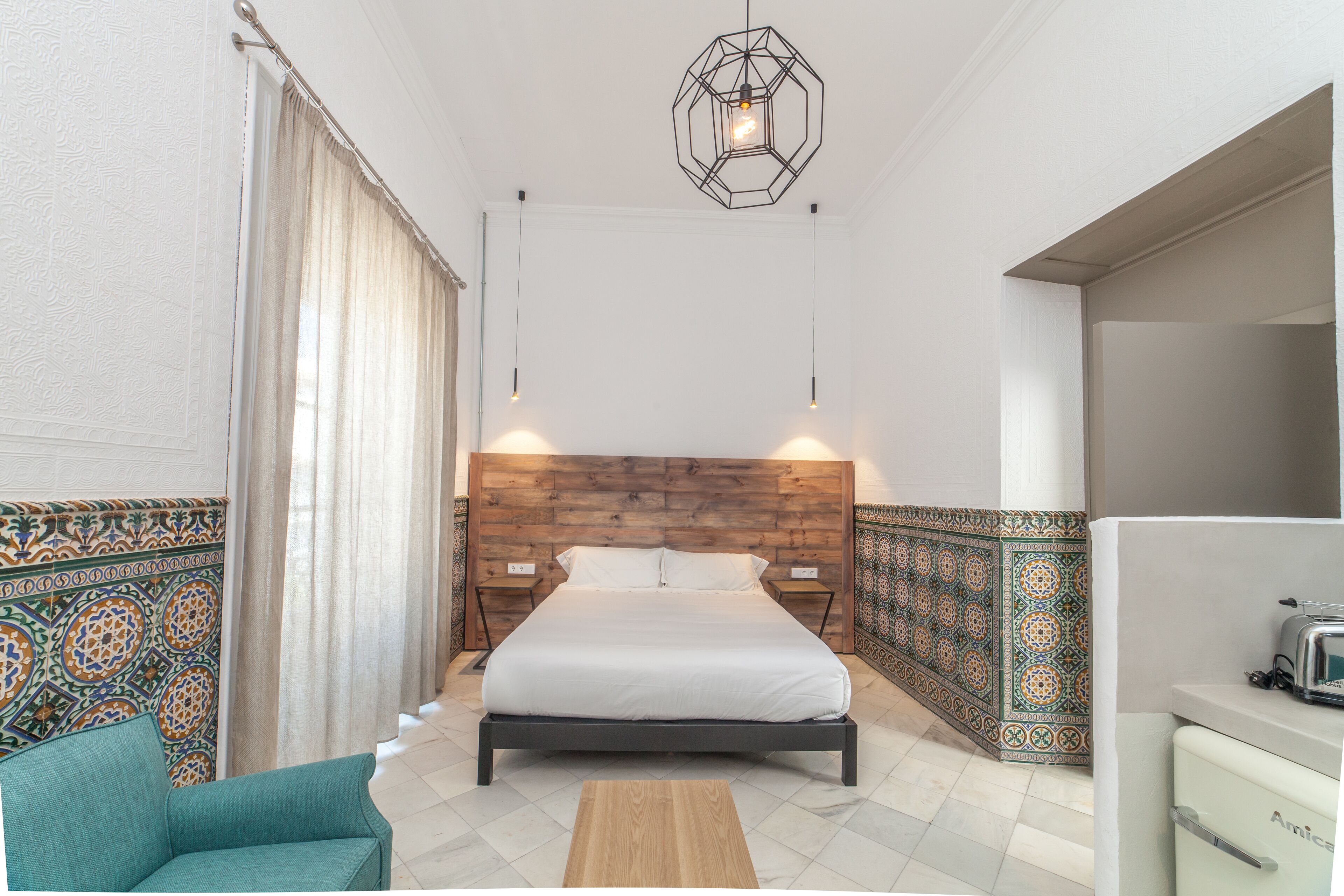 A bedroom with a wooden headboard, a bed, a nightstand, a chair, a wooden coffee table, and a kitchenette with a toaster.