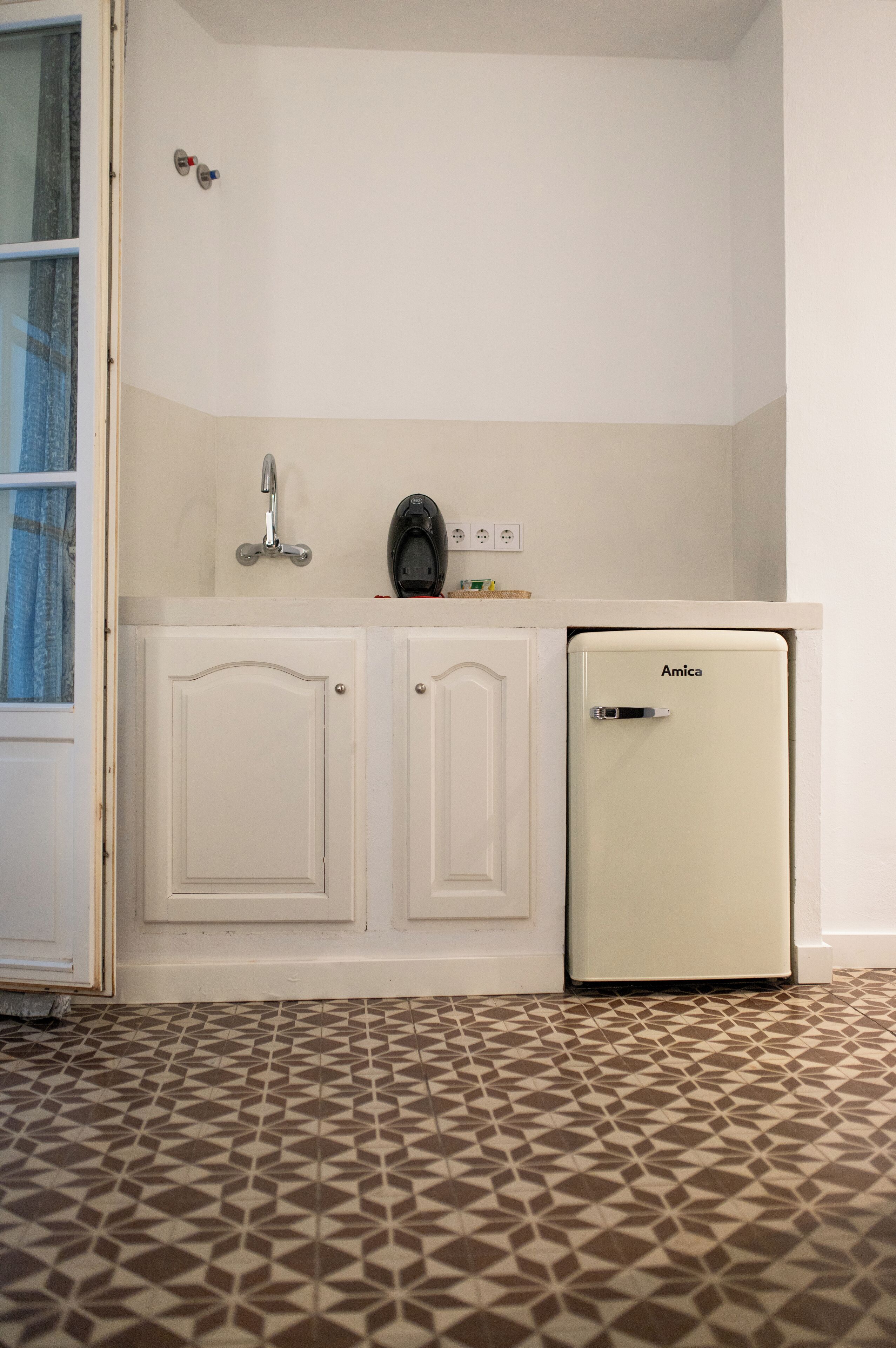 A compact kitchenette with a white cabinet, a sink, and a small refrigerator labeled 'Amica'.