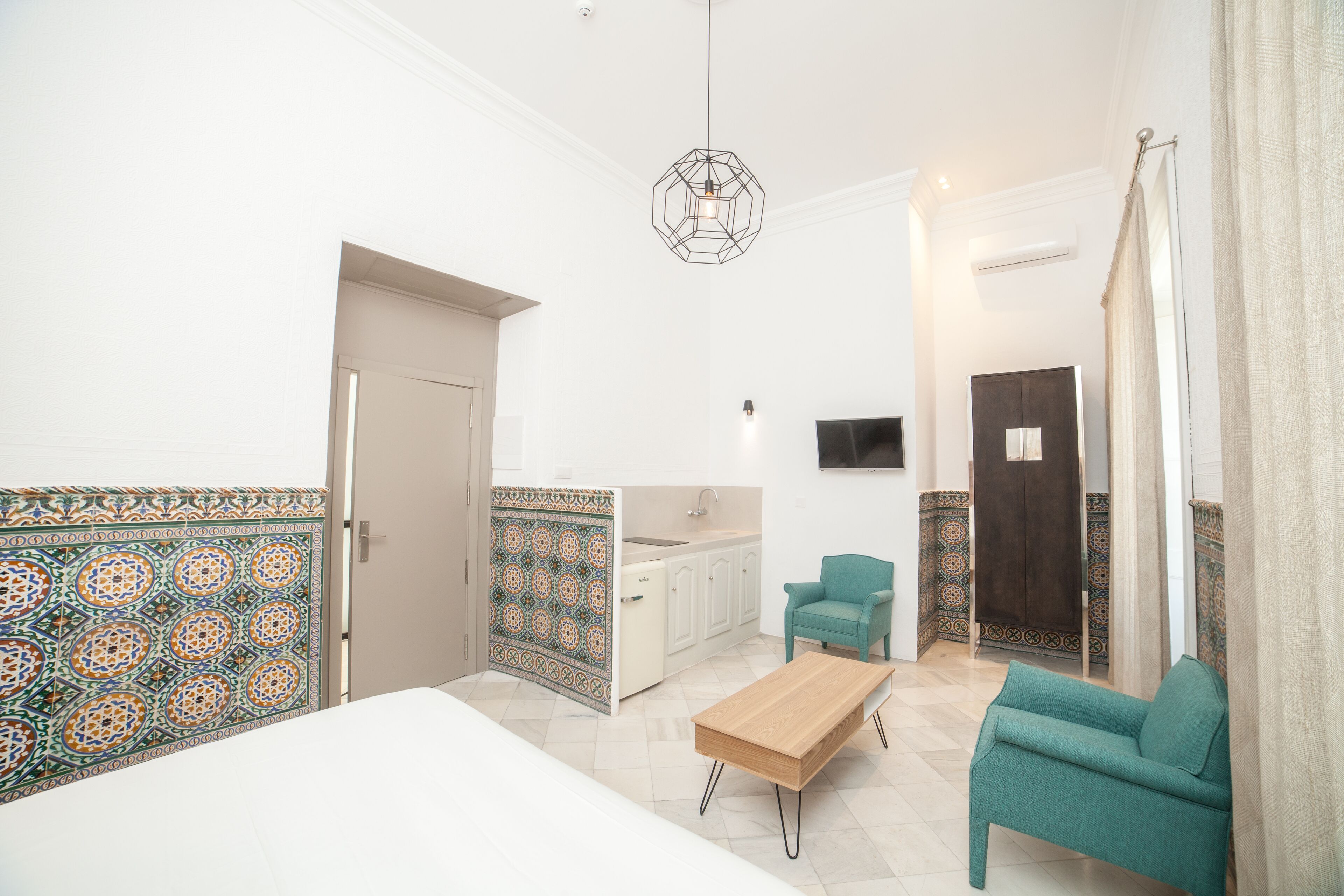 A modern room with a bed, a wooden coffee table, a teal armchair, and a kitchen area with a sink and cabinets.