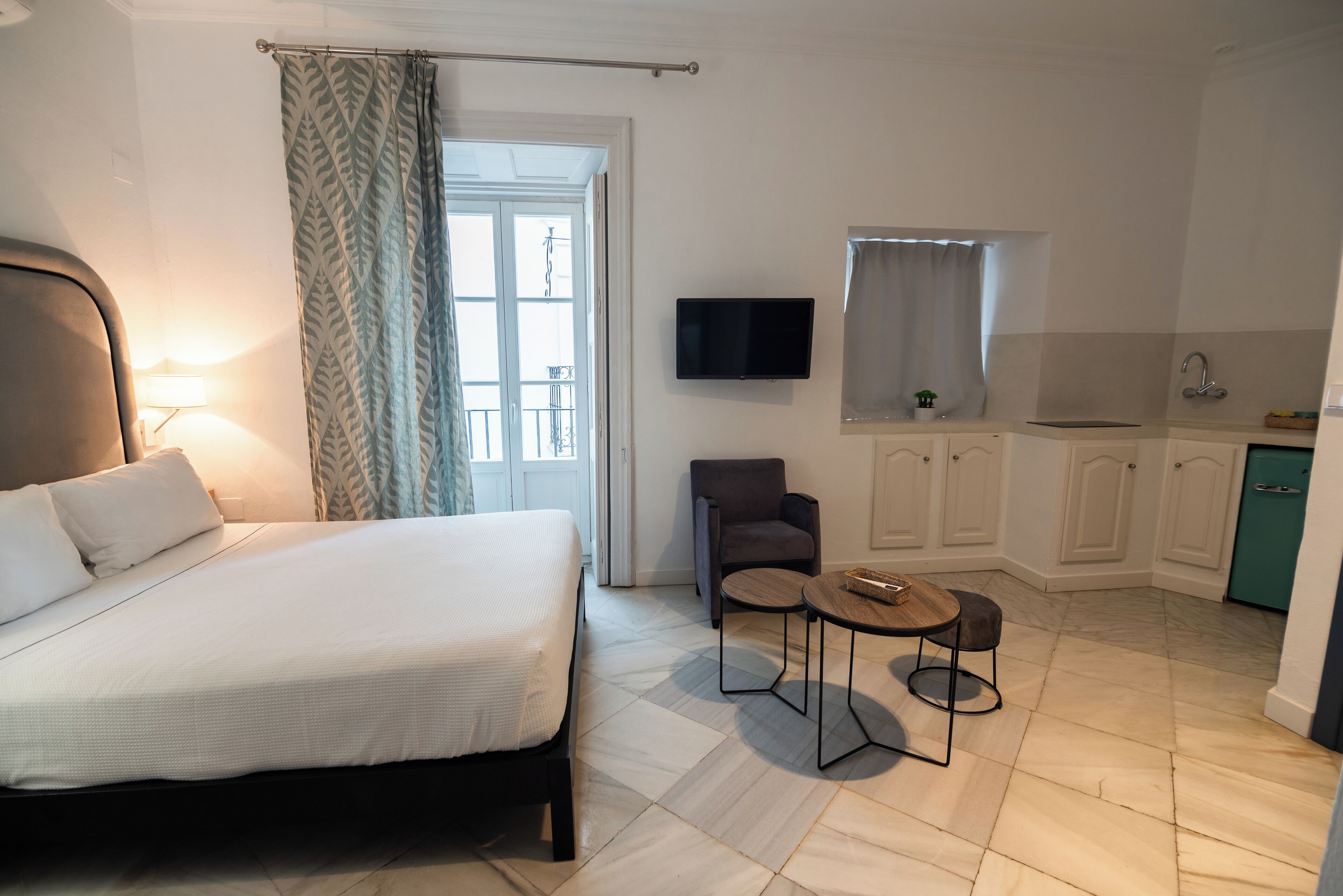A bedroom with a bed, a chair, a small table, a TV, and a kitchenette.