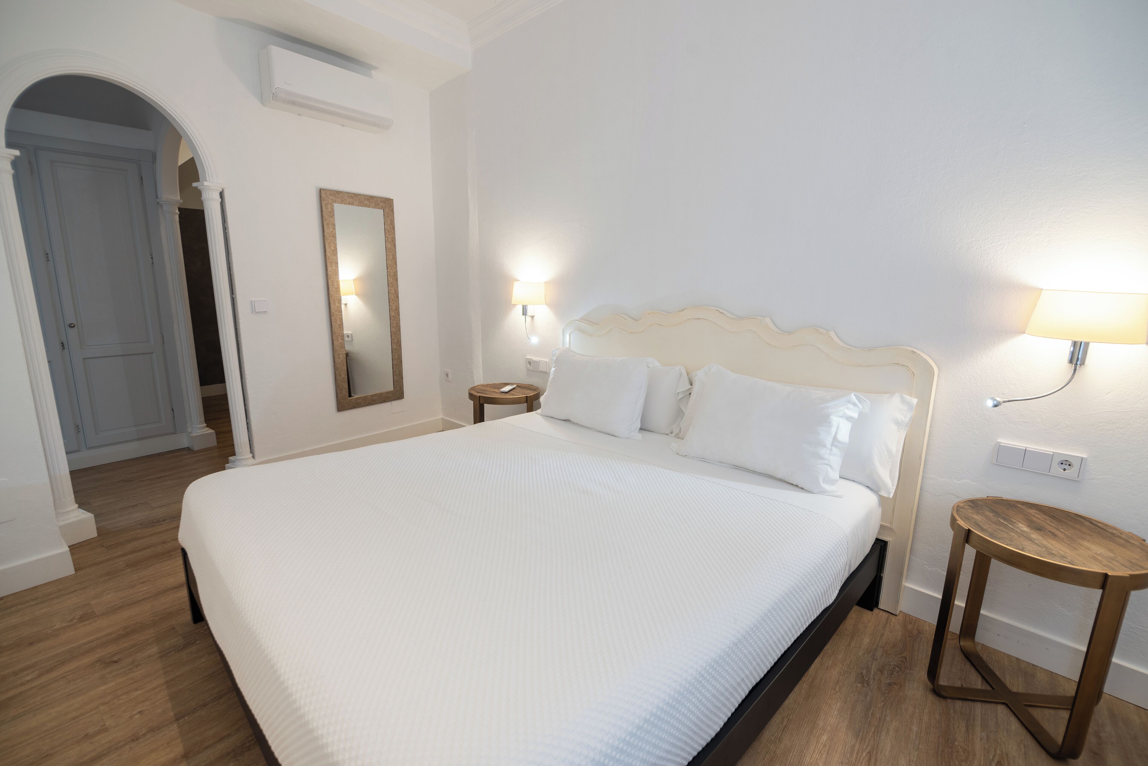 Una cama bien hecha con sábanas blancas en una habitación con suelo de madera, una mesita de noche pequeña y una lámpara de pared.