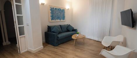 Una sala moderna con un sofá verde oscuro, una mesa de centro de madera y sillas blancas. Un televisor de pantalla plana está montado en la pared, y hay un cuadro sobre el sofá.