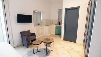 Un espace de vie compact comprenant un canapé, deux petites tables, un téléviseur fixé au mur, un coin cuisine avec des placards et une porte.