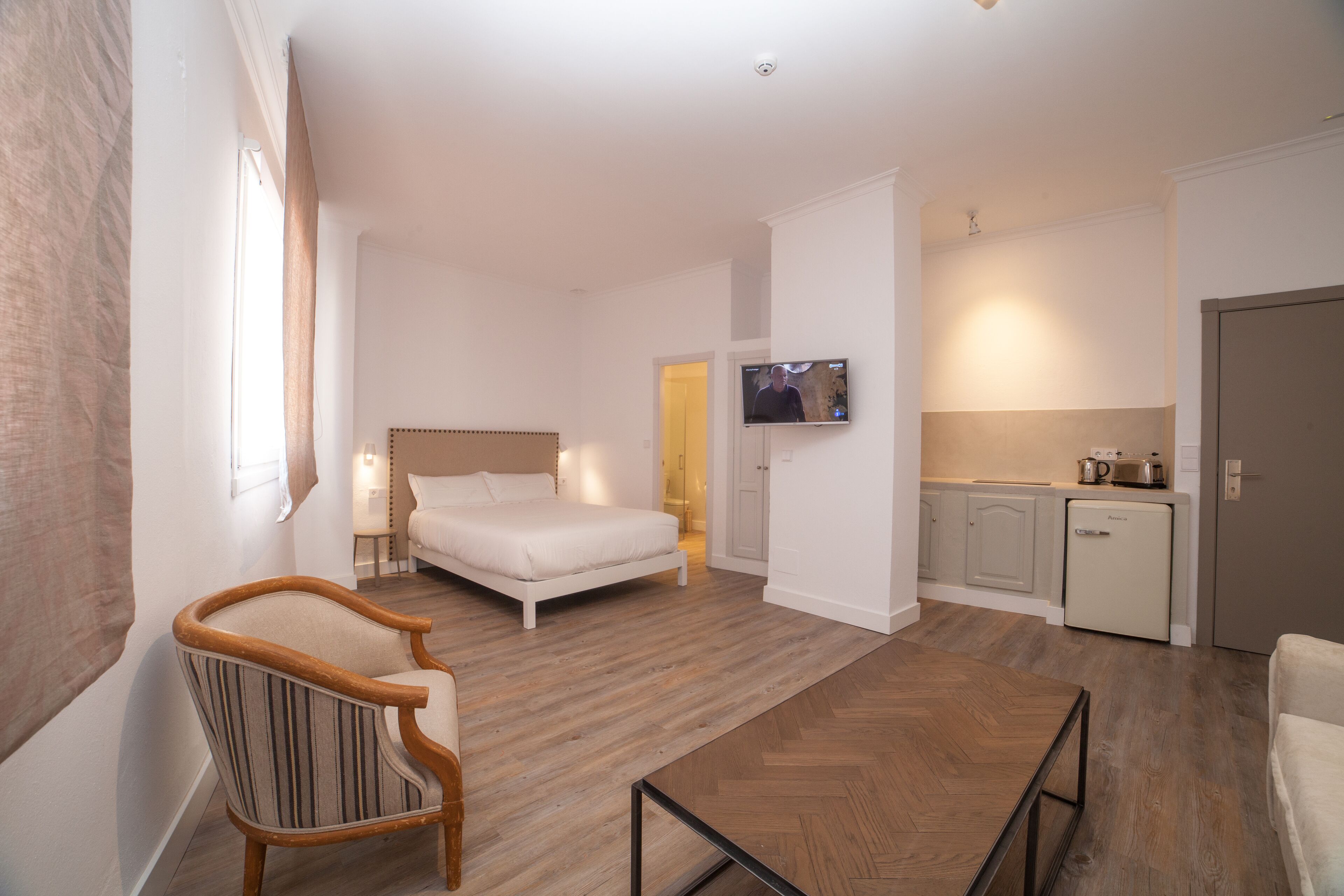 Una habitación de hotel moderna con una cama, una silla, una mesita, un televisor y una kitchenette.