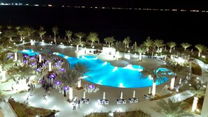 A nighttime view of a resort with a large swimming pool, palm trees, and outdoor seating areas. - Al Bahar Hotel & Resort (Al-Fujairah)