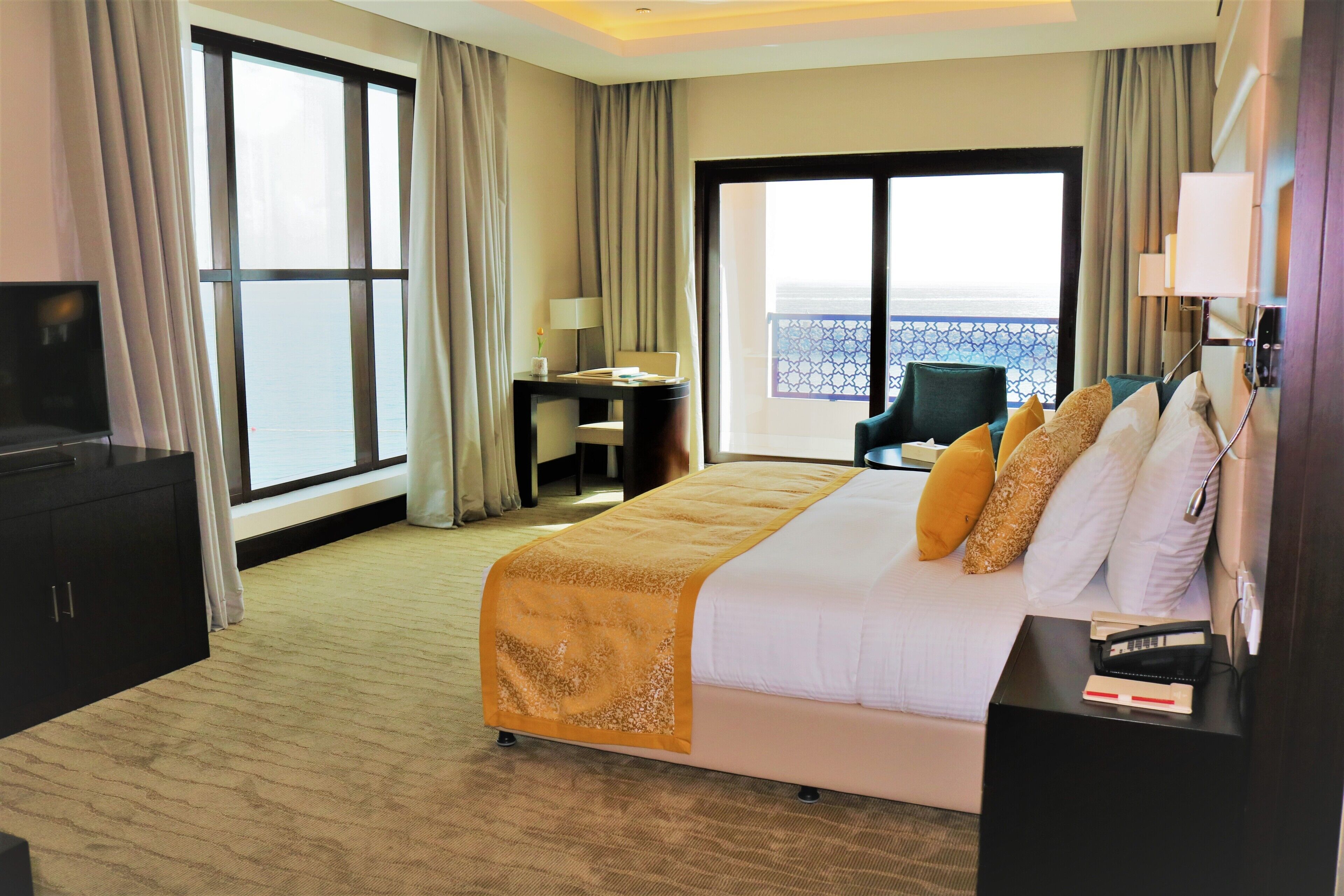 A hotel room with a bed, a desk, a TV, and a view of the sea.